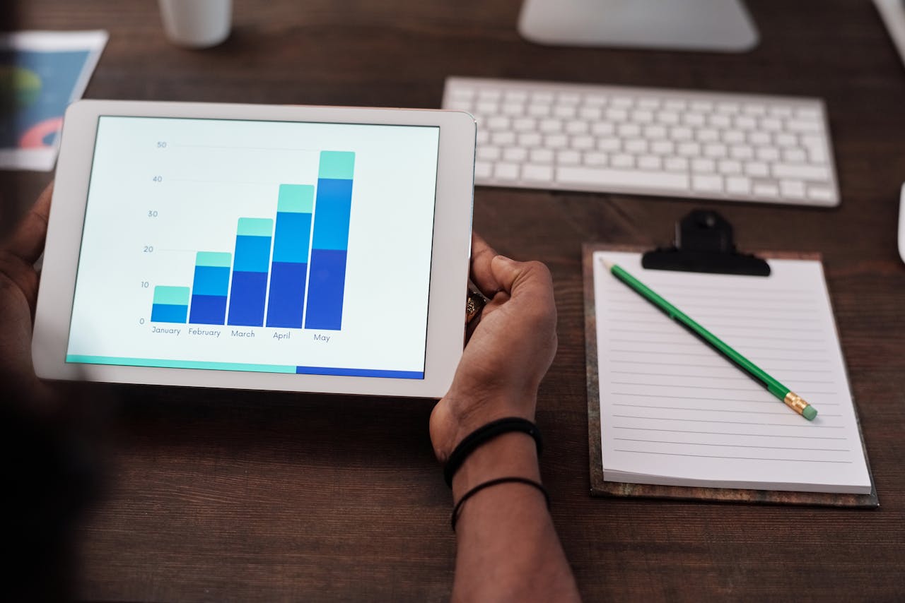 heros-img Close-up of a person holding a tablet showing growth charts on a wooden desk setup.